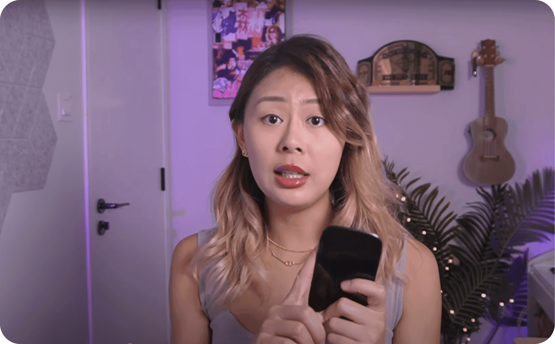 Video still of a woman with long wavy hair holding a black phone-sized device and talking to the camera